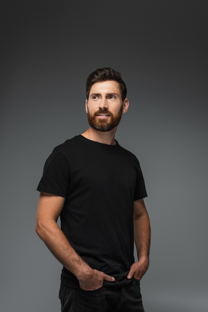 portrait of pleased bearded man in black t-shirt posing with hands in pockets isolated on grey - Fotó, kép