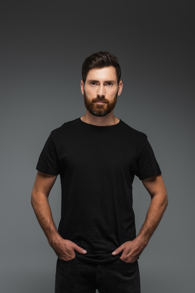 портрет бородатого чоловіка в чорній футболці дивиться на камеру, позує руками в кишенях ізольовані на сірому
 - Фото, зображення