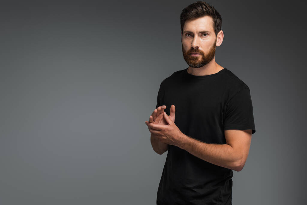 confident man with beard standing in black t-shirt and looking at camera isolated on grey - Foto, Imagen