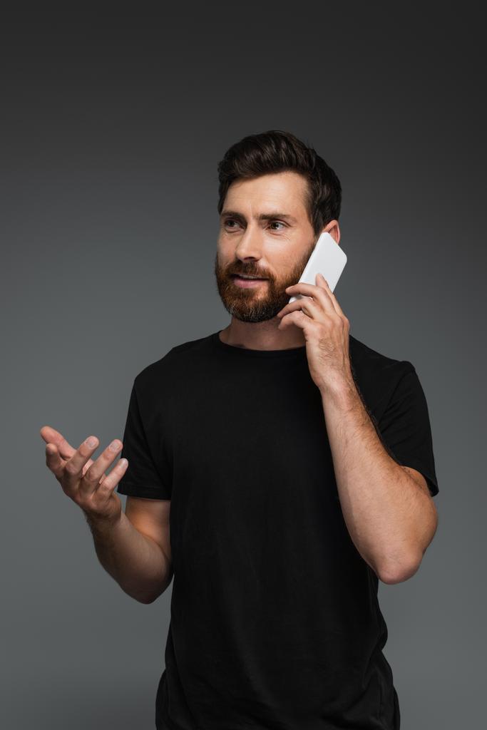 γενειοφόρος άνδρας με μαύρο t-shirt μιλάει σε smartphone απομονωμένο σε γκρι - Φωτογραφία, εικόνα