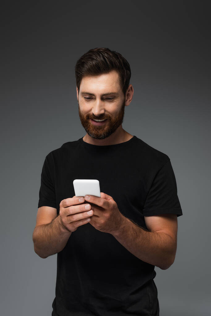 счастливый бородатый мужчина в черной футболке сообщения на смартфоне изолированы на серый - Фото, изображение