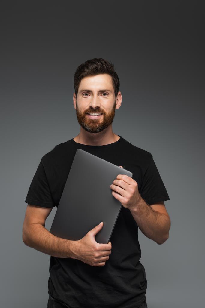 happy bearded man in black t-shirt holding laptop isolated on grey - 写真・画像