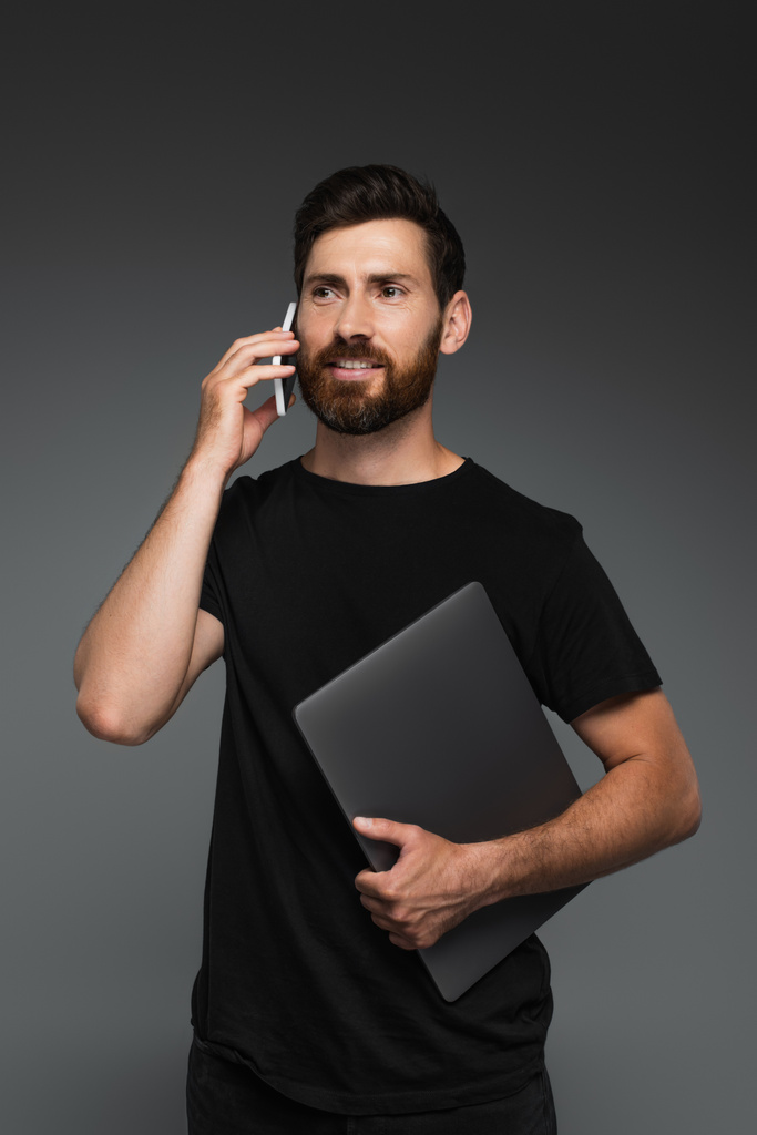 счастливый бородатый мужчина в черной футболке держа ноутбук, разговаривая на смартфоне изолированы на сером - Фото, изображение