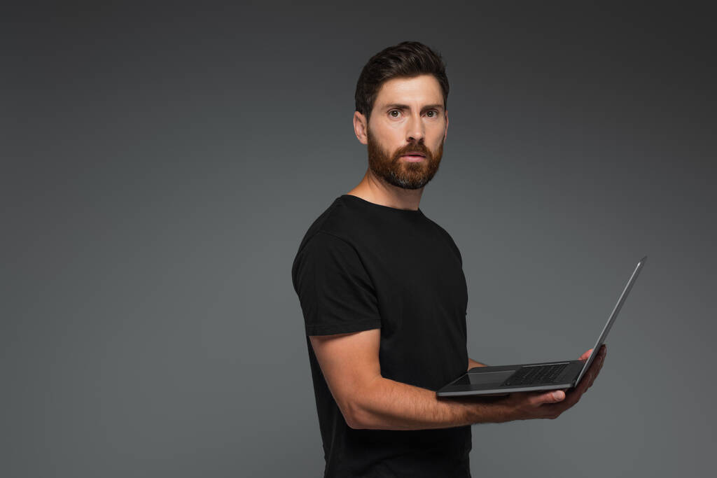 bearded freelancer in black t-shirt holding laptop isolated on grey - Foto, imagen