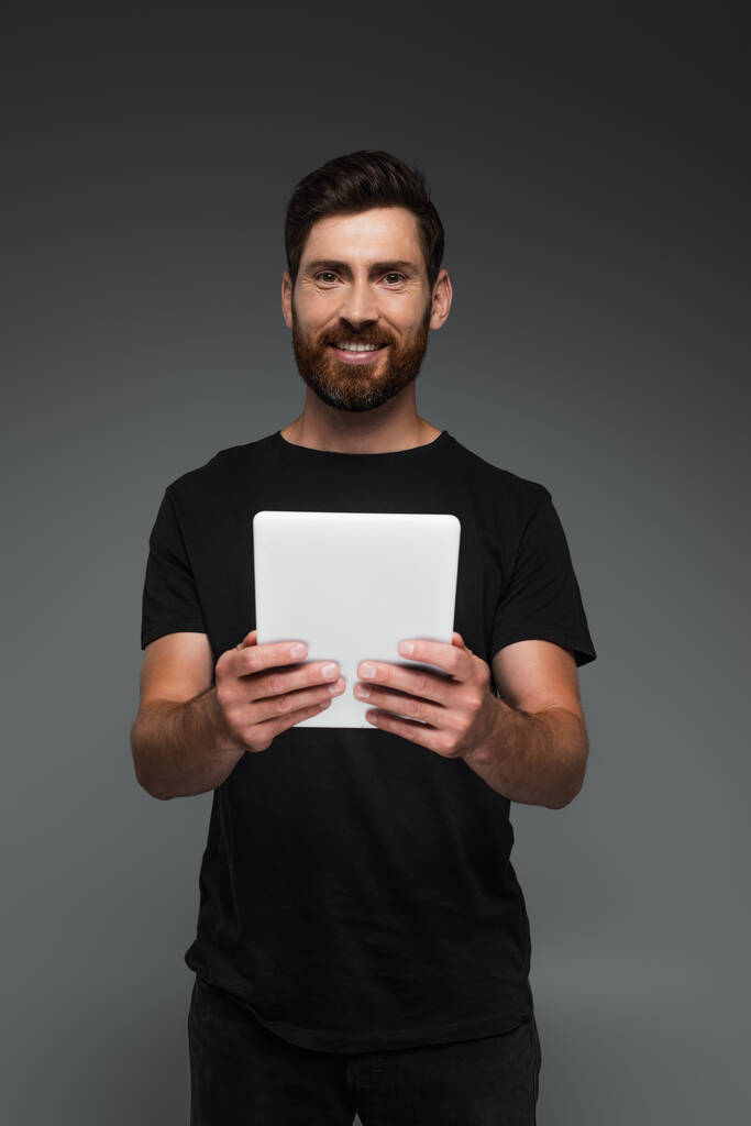 веселий і бородатий чоловік у чорній футболці тримає цифровий планшет ізольований на сірому
 - Фото, зображення