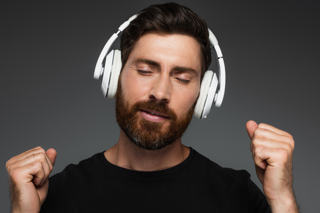 γενειοφόρος άνδρας με κλειστά μάτια ακούει μουσική σε ασύρματα ακουστικά που απομονώνονται σε γκρι - Φωτογραφία, εικόνα