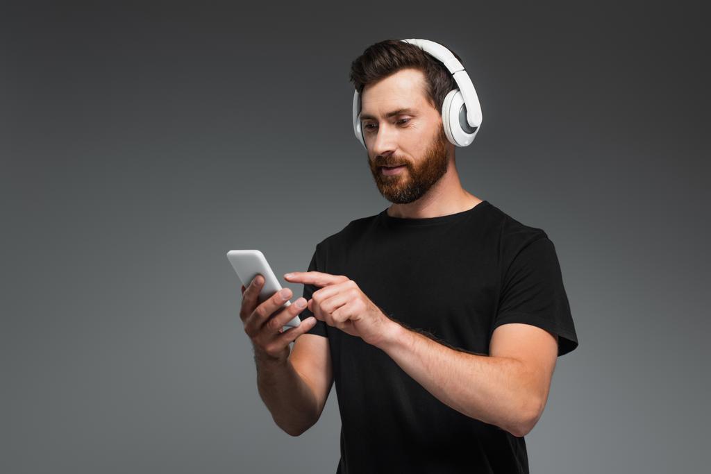 бородатый человек в беспроводных наушниках слушает музыку и указывает на смартфон изолирован на серый - Фото, изображение