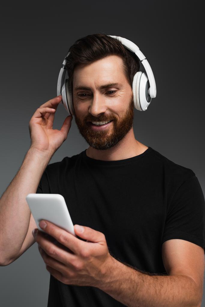 joyful man in wireless headphones listening music and using smartphone isolated on grey - Photo, image