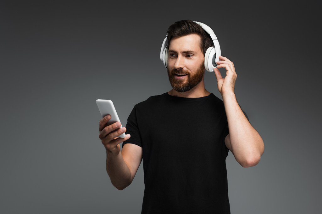 χαμογελαστός άντρας με ασύρματα ακουστικά που ακούει μουσική και χρησιμοποιεί smartphone απομονωμένο σε γκρι - Φωτογραφία, εικόνα