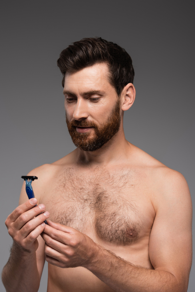 портрет человека без рубашки с бородой, смотрящего на безопасную бритву, изолированную на сером - Фото, изображение