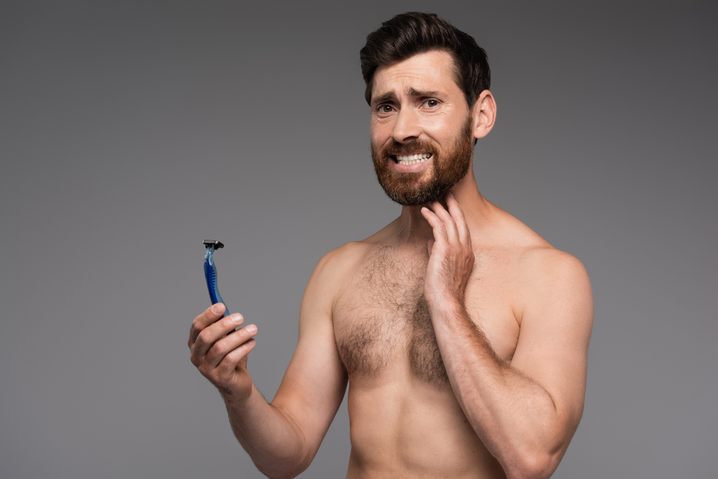 displeased and shirtless man with beard holding safety razor isolated on grey - 写真・画像
