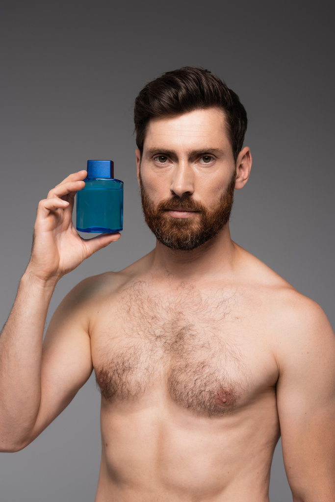 без рубашки человек с бородой держа бутылку с после бритья продукт изолирован на сером - Фото, изображение