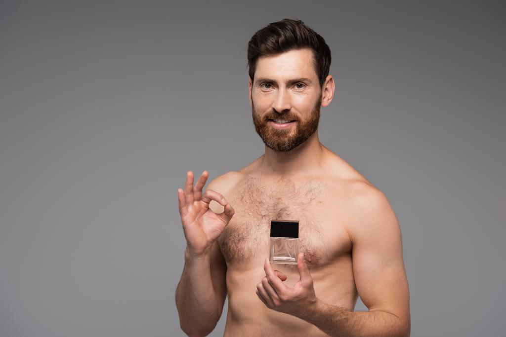 χαρούμενος και shirtless άνθρωπος κρατώντας μπουκάλι με άρωμα και δείχνει ok σημάδι απομονώνονται σε γκρι - Φωτογραφία, εικόνα