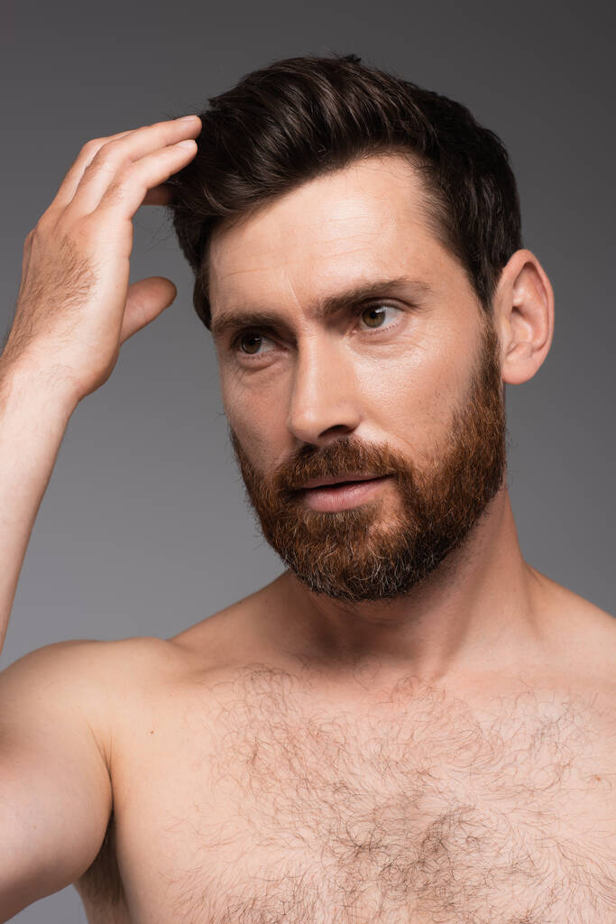 portrait of man with beard adjusting brown hair isolated on grey - Zdjęcie, obraz