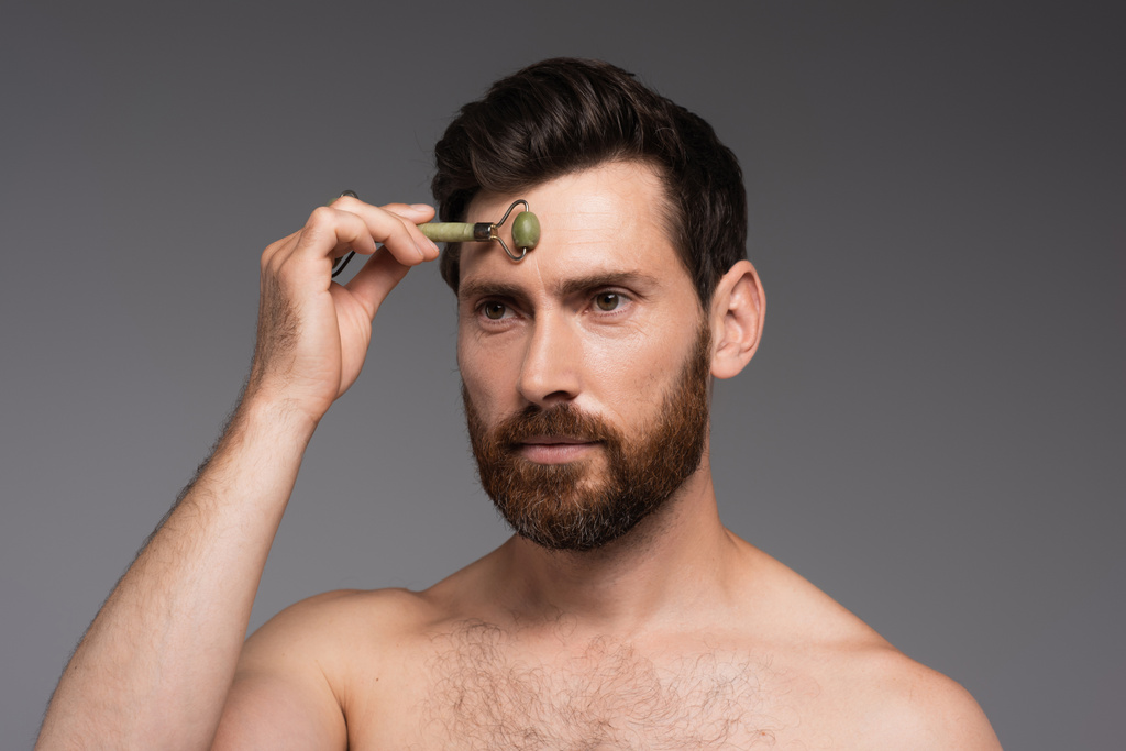 shirtless man with beard using jade roller while massaging forehead isolated on grey - Foto, Imagem