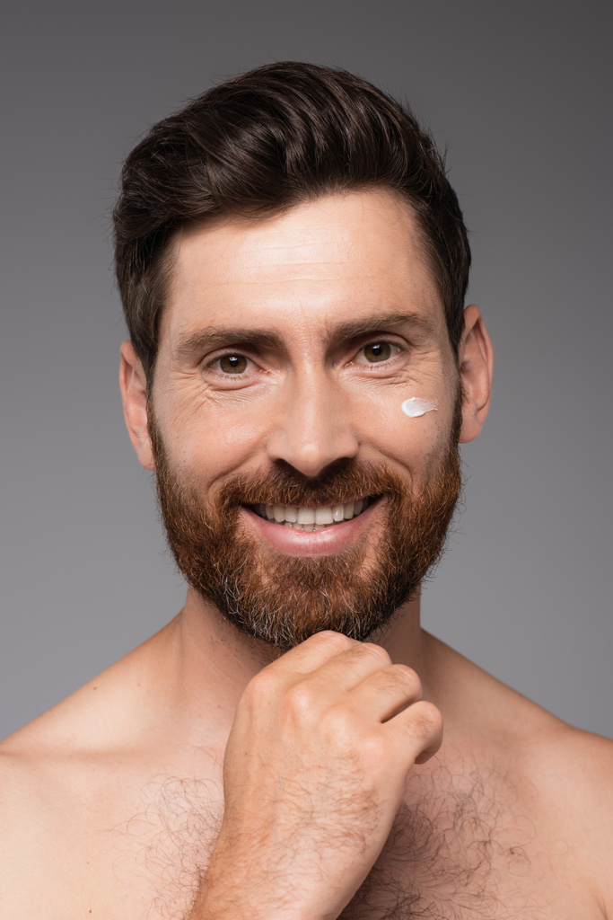 портрет улыбающегося бородатого мужчины со сливками на лице, смотрящего на камеру, изолированную на сером - Фото, изображение