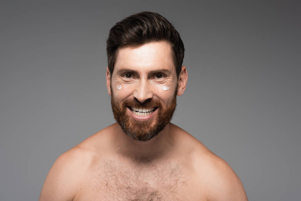 веселый и бородатый мужчина со сливками на лице, улыбающийся изолированно на сером - Фото, изображение