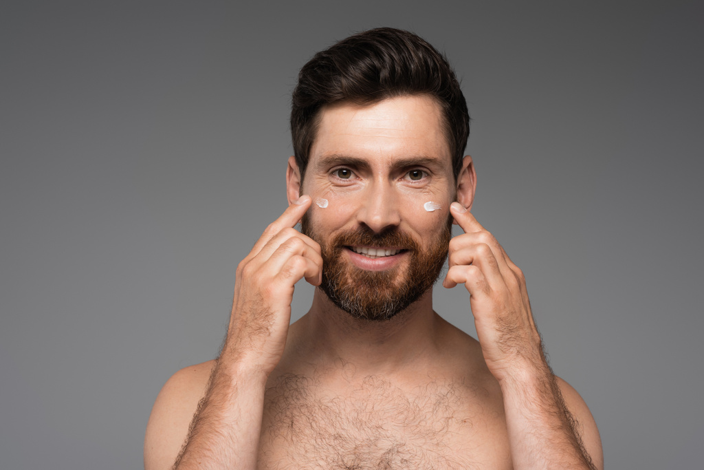 bearded man applying cream on face and smiling isolated on grey - Foto, Imagem