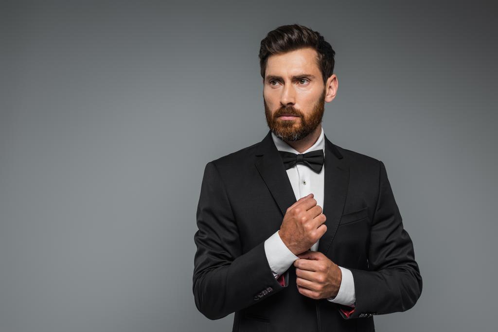 bearded man in black tuxedo with bow tie adjusting sleeve on white shirt isolated on grey - Foto, Imagen
