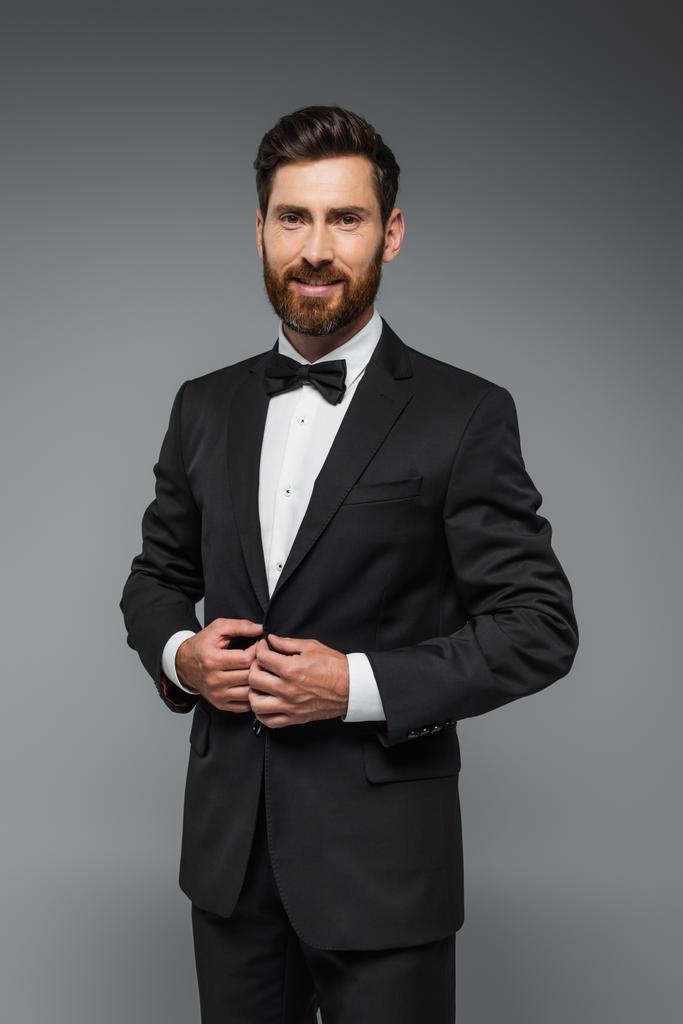 bearded man in black tuxedo with bow tie smiling isolated on grey - Photo, image