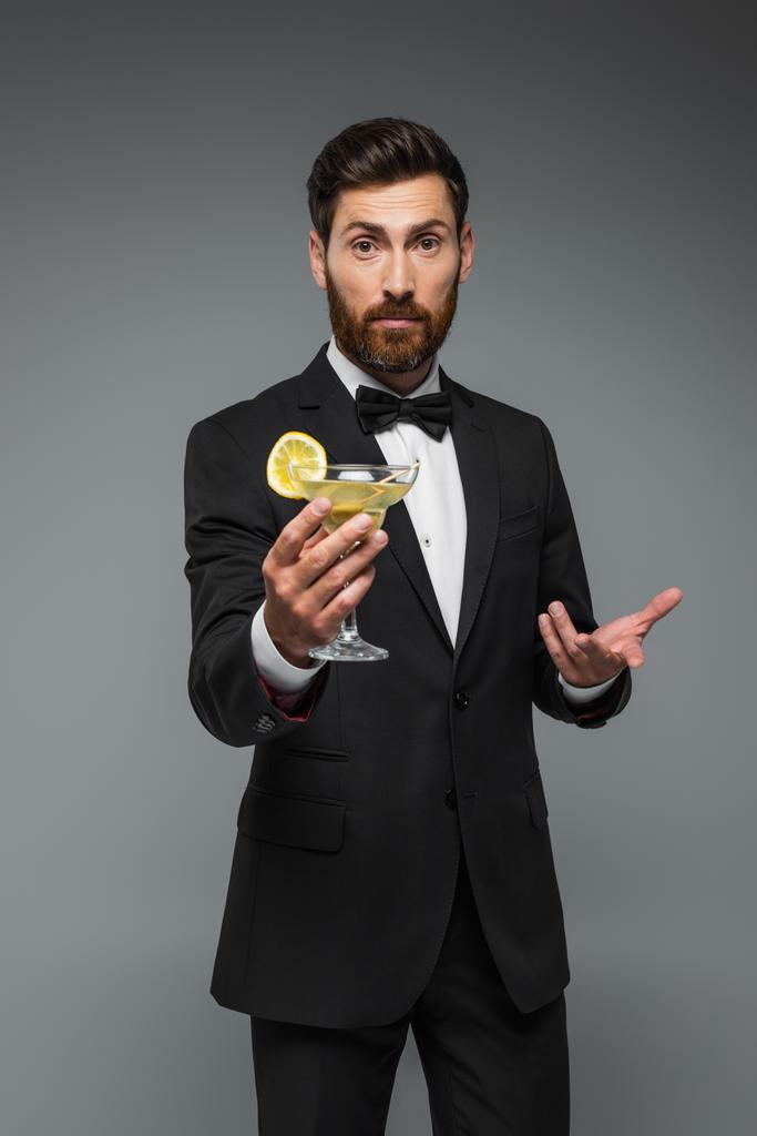 bearded man in elegant suit with holding glass with cocktail and gesturing isolated on grey - Valokuva, kuva