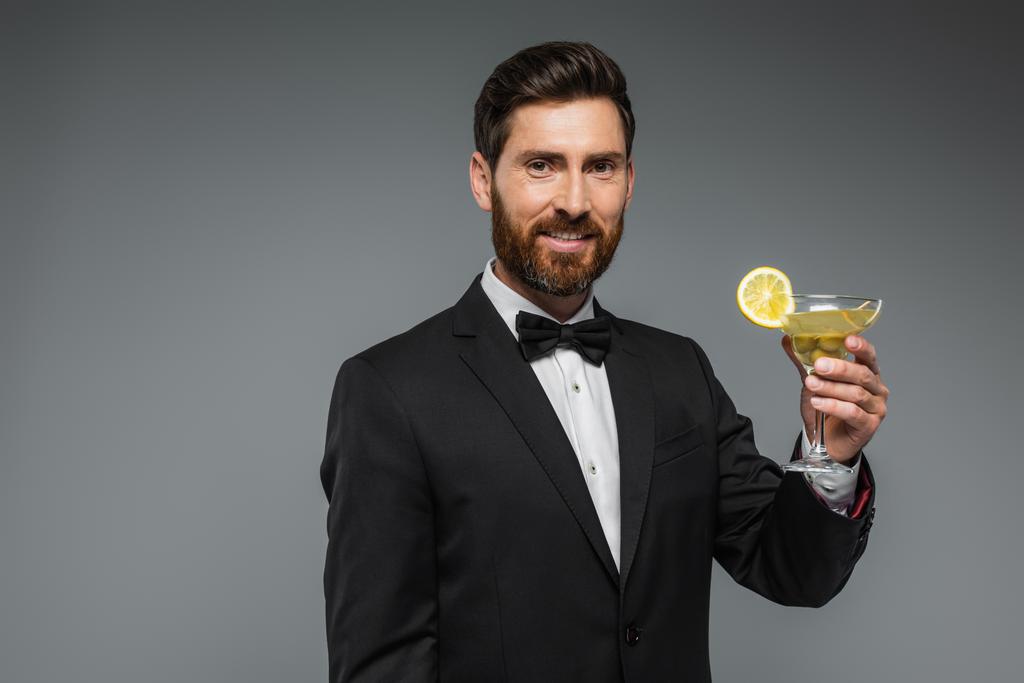 happy man in elegant suit with bow tie holding glass of cocktail isolated on grey - 写真・画像