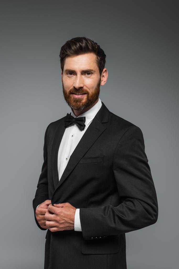 happy and successful man with beard standing in elegant suit with bow tie isolated on grey  - Zdjęcie, obraz