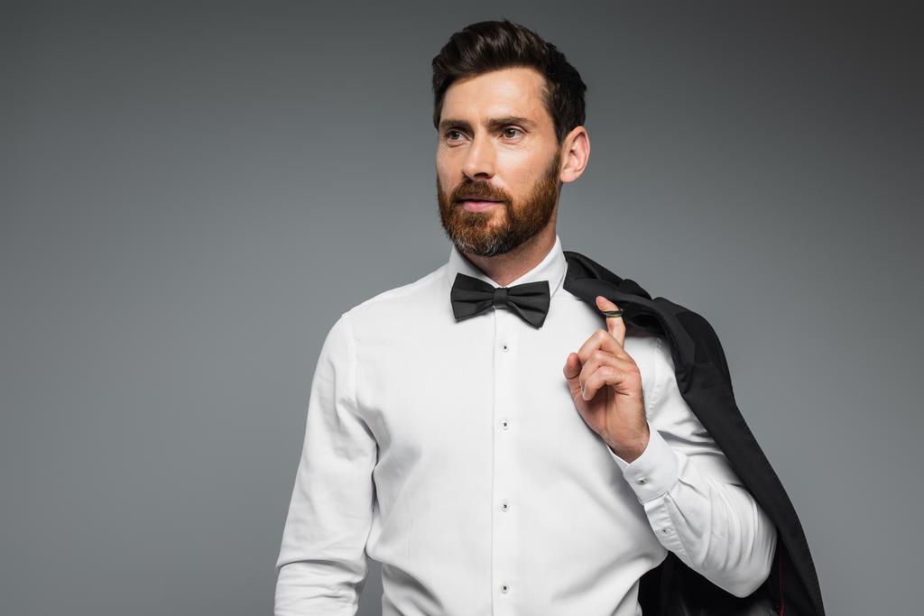 bearded man in white shirt with black bow tie holding blazer isolated on grey  - Foto, Bild
