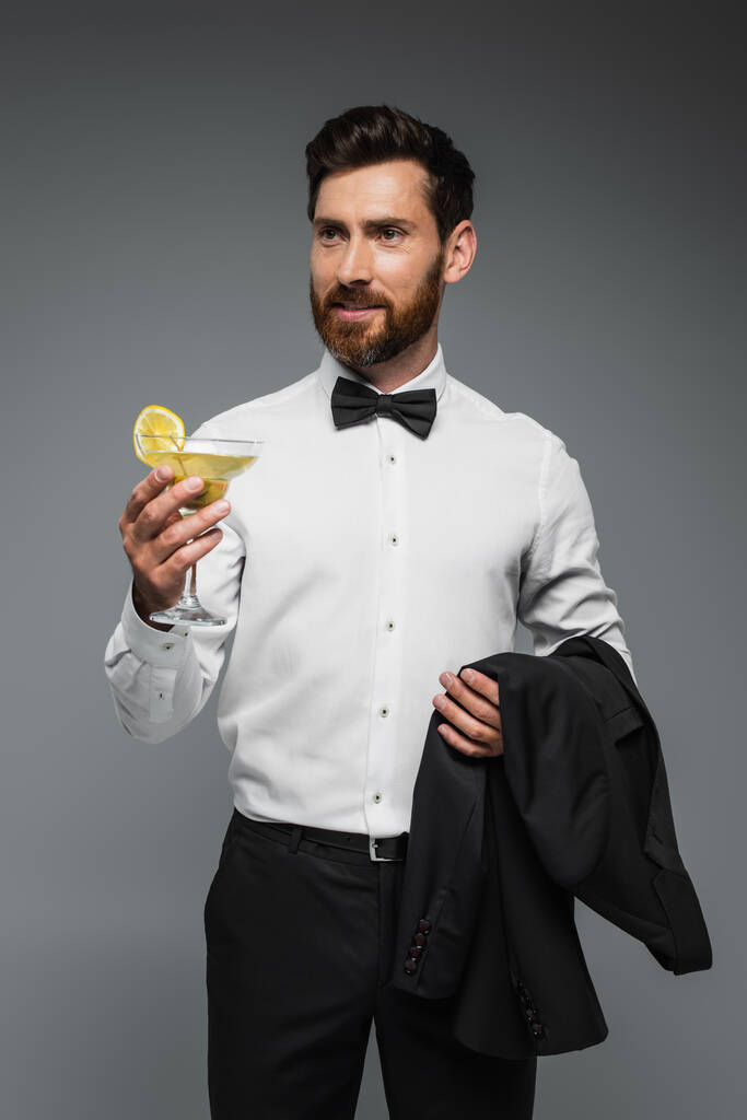 bearded man in tuxedo with bow tie holding glass with cocktail and blazer isolated on grey - Foto, imagen