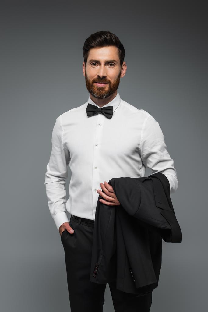 веселый человек в белой рубашке с галстуком-бабочкой держа блейзер и стоя с рукой в кармане изолированы на сером  - Фото, изображение