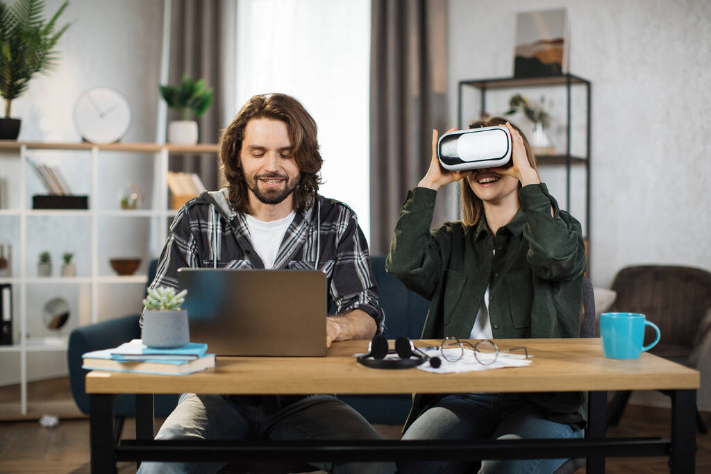 Ευτυχισμένοι νέοι φίλοι που κάθονται στο τραπέζι και χρησιμοποιούν σύγχρονα gadgets για δουλειά ή μελέτη. Όμορφο κορίτσι φοράει γυαλιά εικονικής πραγματικότητας και απολαμβάνει την τεχνητή πραγματικότητα, ενώ εστιασμένος άντρας πληκτρολογεί στο laptop. - Φωτογραφία, εικόνα