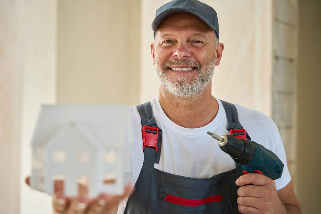 Photo of adult builder in baseball cap and overalls holding small house model in one hand and drill in other - Valokuva, kuva