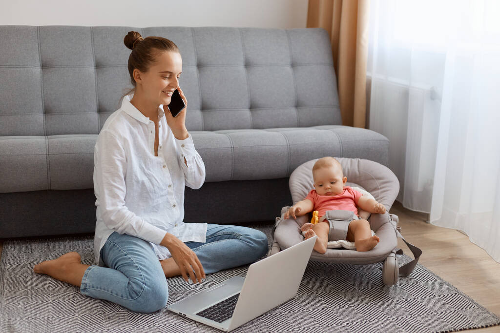 Smiling positive woman in white t shirt sitting near cough with baby daughter in rocking chair and working on laptop, talking on mobile phone, being in good mood. - Fotó, kép