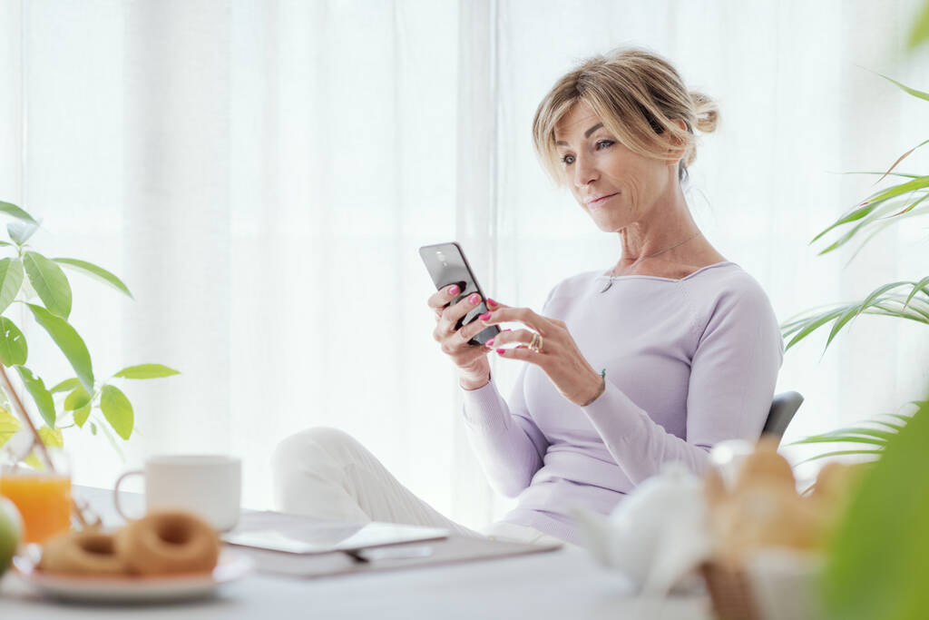 Mature woman having breakfast and connecting with her smartphone at home - Foto, immagini
