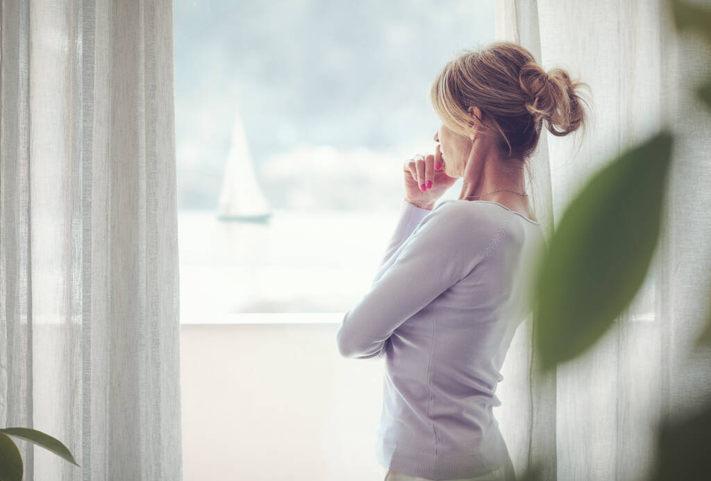 Зрелая женщина расслабляется дома и стоит рядом с окном, смотрит в сторону и думает: - Фото, изображение
