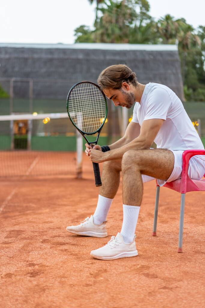 Καυκάσιος ξανθός μακρυμάλλης νεαρός που κάθεται σε μια καρέκλα κρατώντας τη ρακέτα του τένις στο κεφάλι του με μια στάση ήττας σε ένα βρώμικο γήπεδο. - Φωτογραφία, εικόνα