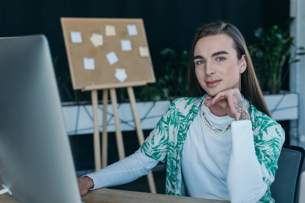 Tattooed queer designer looking at camera near computer in creative agency  - Foto, imagen