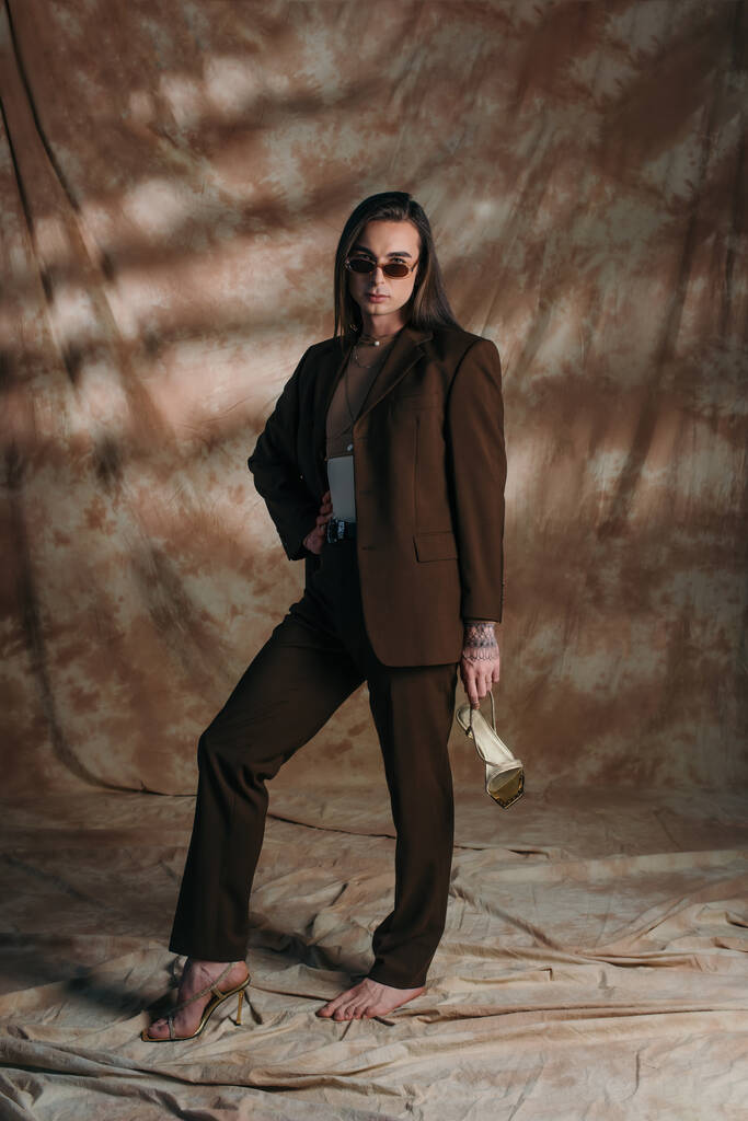 Trendy nonbinary person in suit holding shoe on abstract brown background - Foto, Imagem