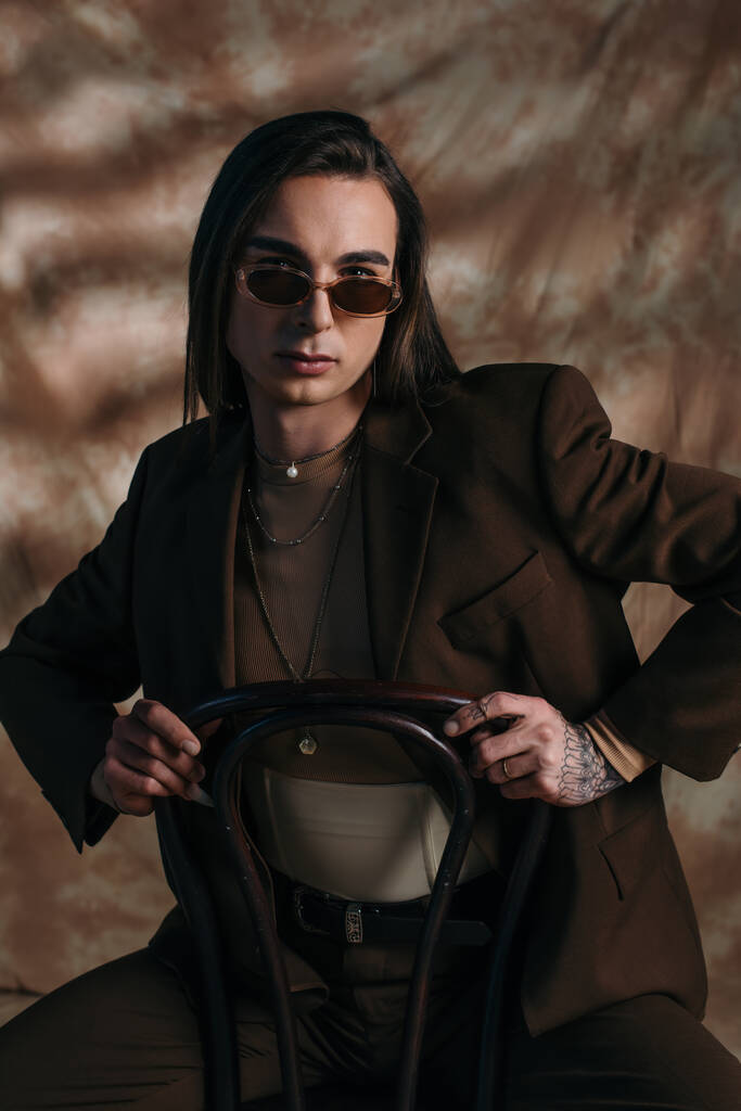 Портрет брюнетки в солнцезащитных очках и куртке, сидящей на стуле на абстрактном коричневом фоне - Фото, изображение