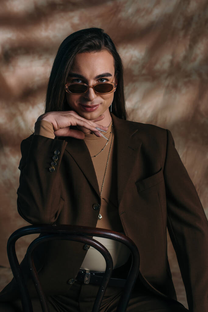 Портрет улыбающегося странного человека в солнцезащитных очках и куртке, смотрящего в камеру, сидя на стуле на абстрактном коричневом фоне - Фото, изображение
