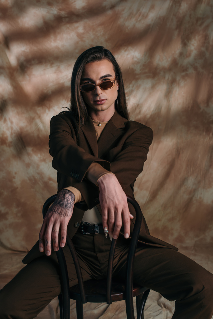 Fashionable tattooed queer person in sunglasses sitting on chair on abstract brown background - Foto, imagen