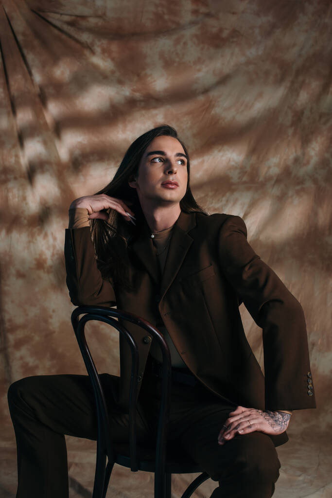 Модный педик в костюме трогает волосы, сидя на стуле на абстрактном коричневом фоне - Фото, изображение