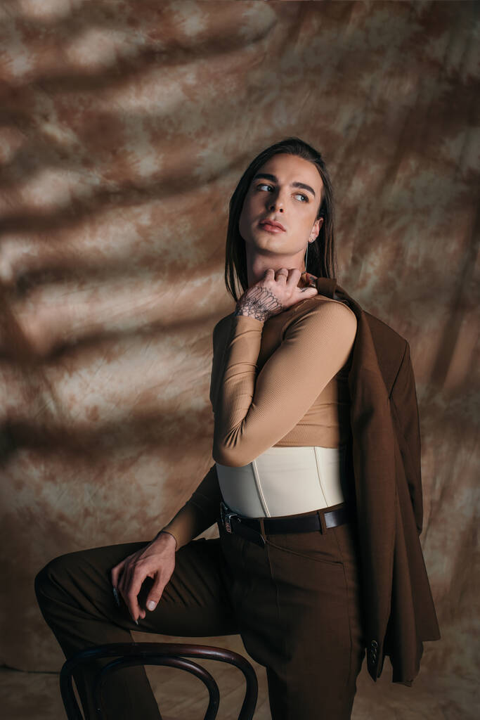 Nonbinary person in corset holding jacket near chair on abstract brown background - Foto, Imagem