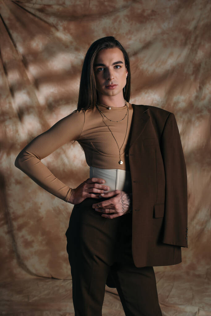 Brunette queer person in jacket holding hand on hip on abstract brown background - Photo, image