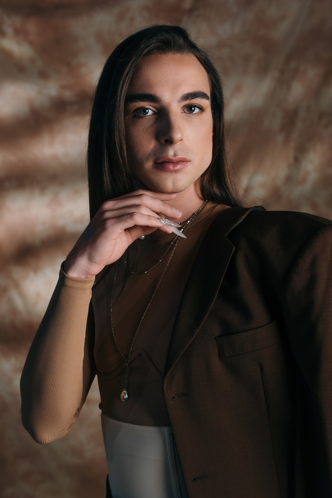 Portrait of stylish nonbinary person touching chin and looking at camera on abstract brown background - Photo, Image