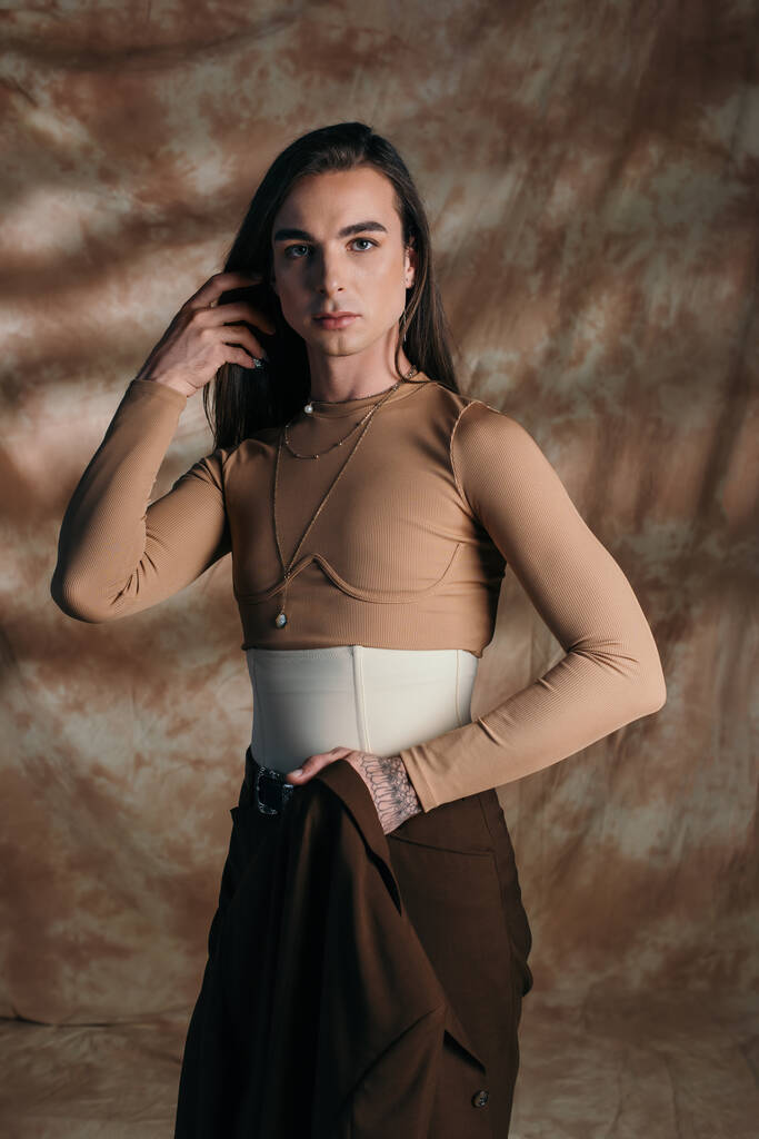 Stylish nonbinary person holding jacket and looking at camera on abstract brown background - 写真・画像