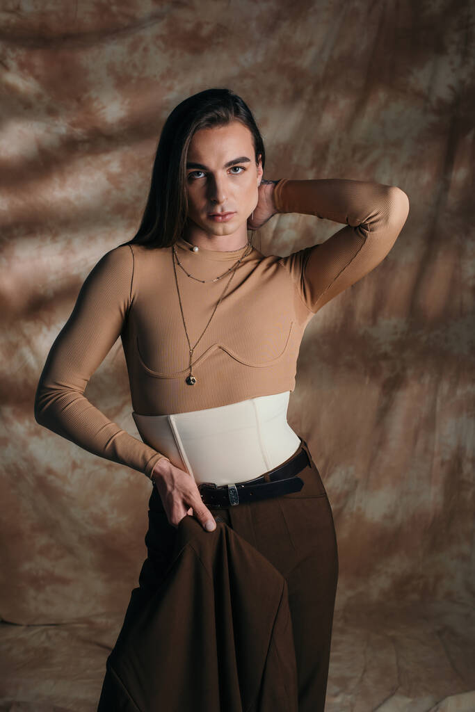 Trendy nonbinary person holding blazer and posing on abstract brown background - Fotoğraf, Görsel