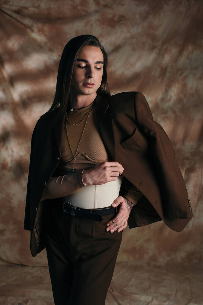 Stylish brunette queer person touching jacket on abstract brown background - Fotografie, Obrázek