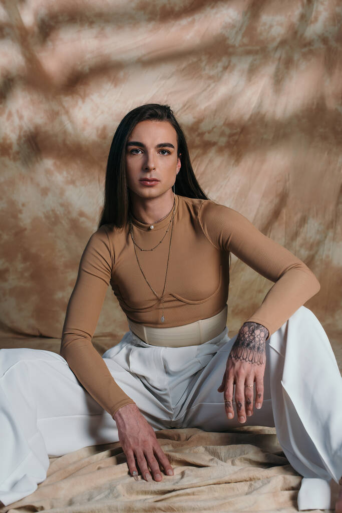 Fashionable queer person in corset and white pants looking at camera on abstract brown background  - Foto, afbeelding