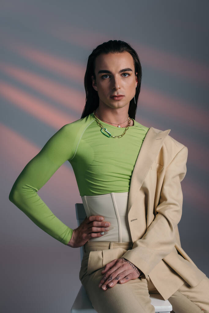 Trendy νεαρό queer μοντέλο στο κοστούμι και κορσέ ποζάρουν σε καρέκλα σε αφηρημένο φόντο  - Φωτογραφία, εικόνα